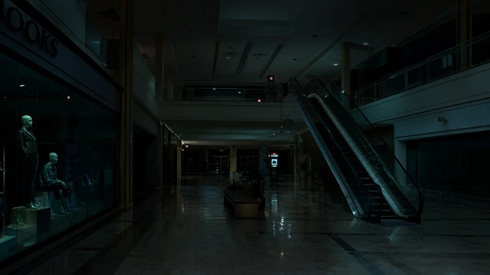 An eerie, empty shopping mall at night with non-moving escalators and dark shop fronts, creating a sense of social absence.