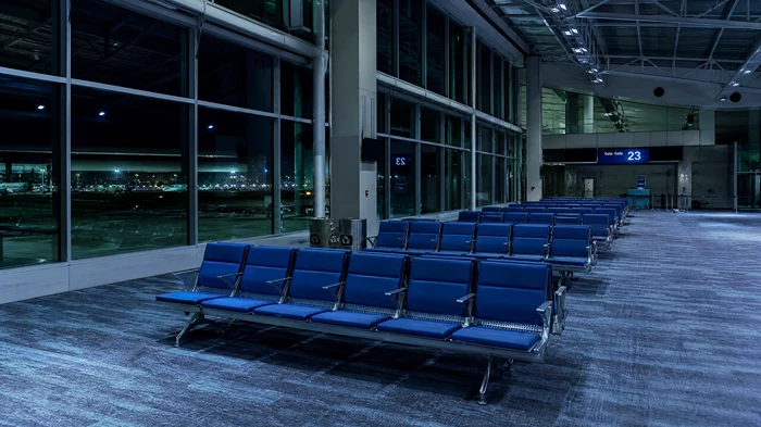 A vast and empty airport terminal at night with rows of blue seats, cold blue lighting, and large windows looking out onto a dark runway.